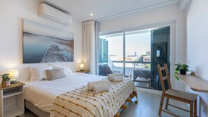 Ein Hotelzimmer mit Bett, Sessel, Tisch, Lampe, einem Gemälde und einem Balkon mit Blick.