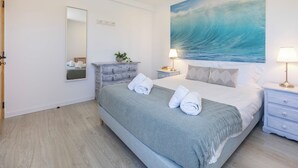 Ein Hotelzimmer mit einem Bett, einem Schminktisch, einem Spiegel, einem Gemälde einer Welle und einem Nachttisch mit Lampe.