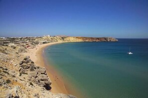 A coastal landscape with a sandy beach, rocky cliffs, and a clear blue sea.