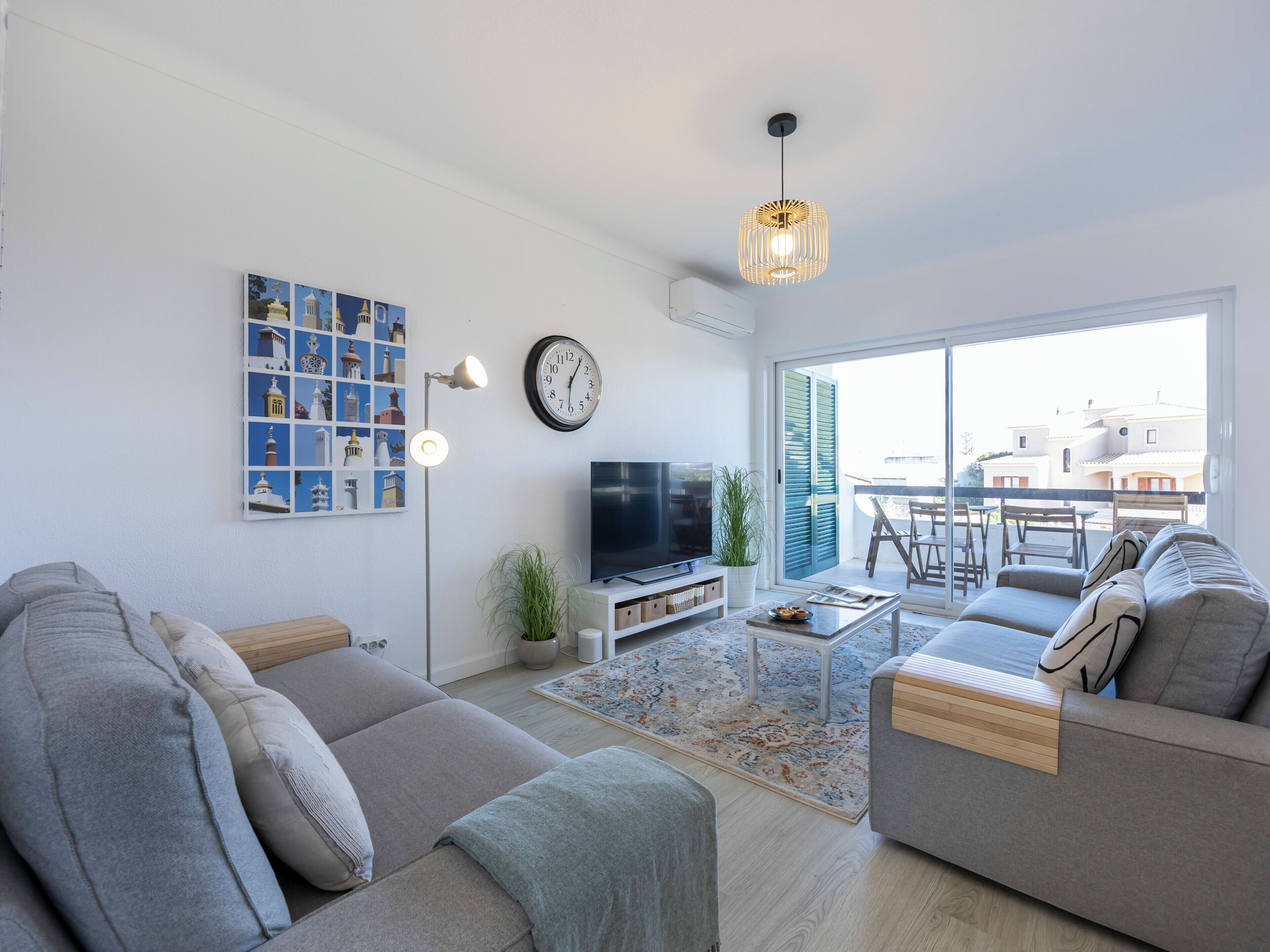 A modern living room with a sofa set, a flat-screen TV, a clock, and a large window offering a view of buildings.