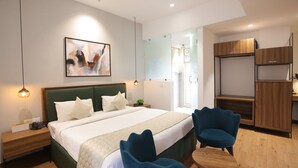 A modern bedroom with a large bed, two blue chairs, a wooden desk, and a wardrobe. - Pondy Heights - Suites and Studios (Puducherry)