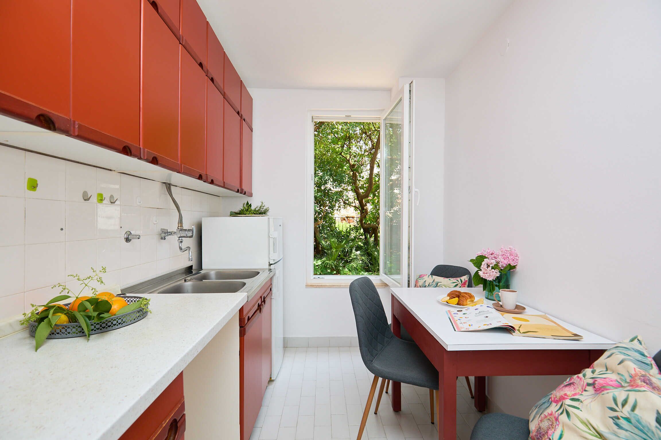 A compact kitchen with red cabinets, a white countertop, and a window overlooking greenery.