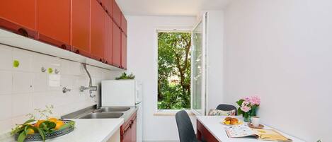 A compact kitchen with red cabinets, a white countertop, and a window overlooking greenery.