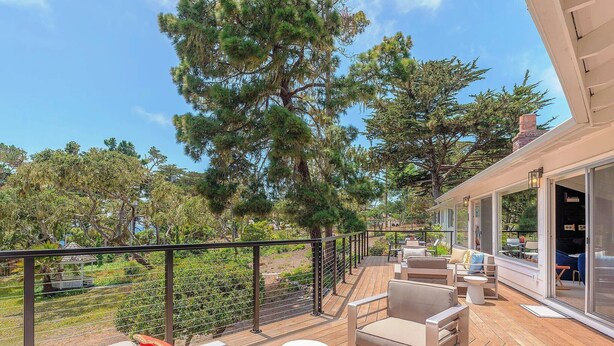 Une terrasse en bois, dotée de meubles d’extérieur, d’une rambarde et offrant une vue sur des arbres et un bâtiment.