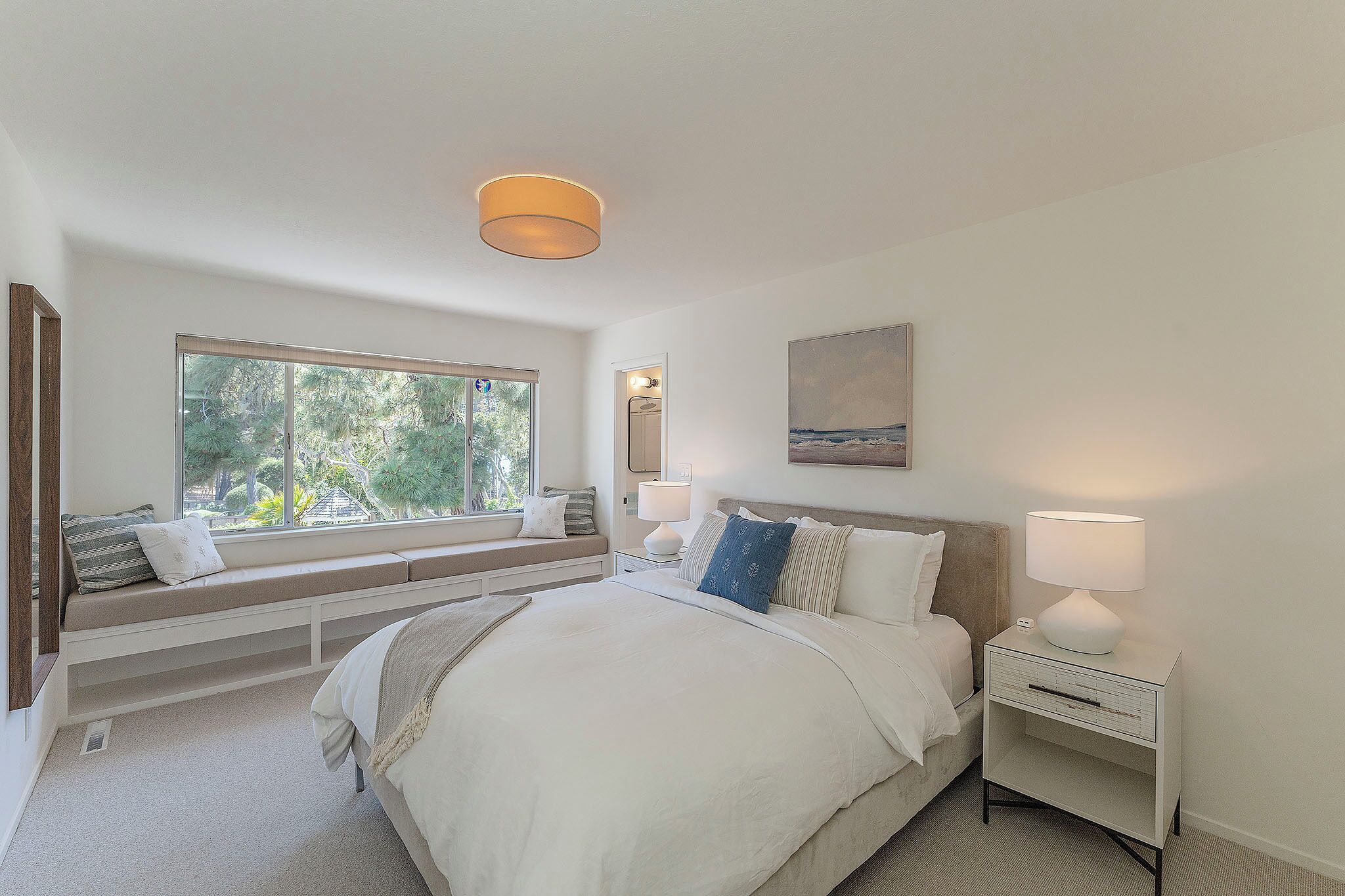 A bedroom with a large bed, a window seat, a bedside table with a lamp, and a painting on the wall.