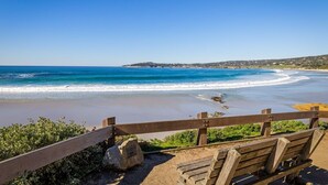 Un banc en bois donnant sur une plage où l’eau est d’un bleu limpide, avec une côte visible au loin.