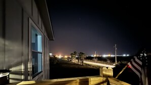 A nighttime view from a balcony with a wooden railing, overlooking a lit area with palm trees and a distant building. - Relax & Unwind in a Serene Retreat Near the Beach (Port Arthur)
