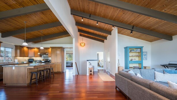 Una cocina amplia con armarios de madera y una isla grande, un espacio de sala acogedor con un sofá y un armario azul, y un techo alto con vigas a la vista.