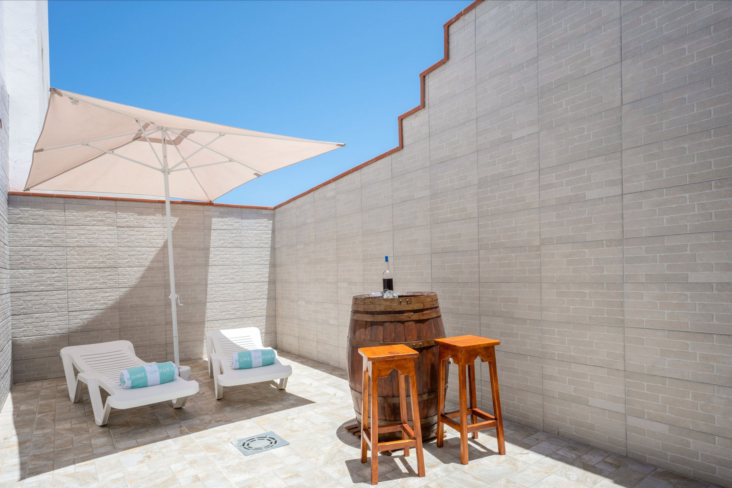 Un espace patio agrémenté d’un parasol blanc, de deux tabourets en bois et de deux chaises longues blanches avec des coussins bleus.