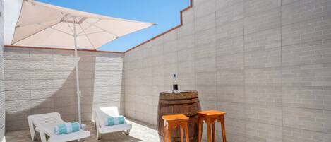 A patio area with a white umbrella, two wooden stools, and two white lounge chairs with blue cushions.