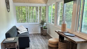 Una sala de estar acogedora con un sofá gris oscuro, una mesa de centro de madera y un televisor sobre un mueble de madera. Amplias ventanas ofrecen vistas a un área boscosa.