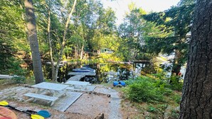 Un área boscosa con un muelle que se adentra en un lago tranquilo, rodeado de árboles y una mesa de picnic junto a kayaks.