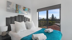 A modern hotel room with a large bed, white bedding, a teal throw blanket, and a view of a cityscape through a large window. - Atlantic Port 9 (Cape Town)