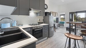 A modern kitchen with grey cabinets, a stainless steel refrigerator, a gas stove, and a sink. There is a dining area with a table and chairs visible through an open door. - Atlantic Port 9 (Cape Town)