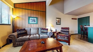 Sala de estar con sofá, sillón y mesa de centro. Hay dos cuadros enmarcados en la pared, una lámpara y una ventana con persianas.
