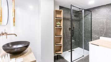 Ein modernes Badezimmer mit einem schwarzen Waschbecken, Holzregalen, einer Duschkabine aus Glas und Marmorböden.