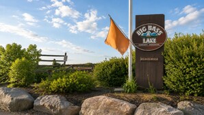 A sign for Big Bass Lake Community Association with a flag, rocks, and greenery. - Family-Friendly Cozy Cabin Hot Tub, Fire Pit, Outdoor Pool & Ping Pong table (Clifton township)