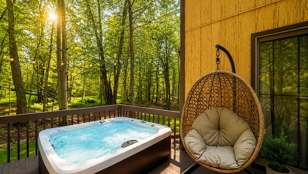 A hot tub on a wooden deck with a hanging chair, surrounded by a forest. - Family-Friendly Cozy Cabin Hot Tub, Fire Pit, Outdoor Pool & Ping Pong table (Clifton township)
