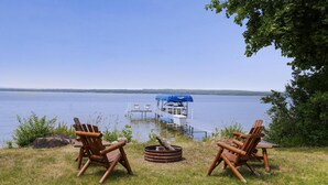 Eine Uferlandschaft mit zwei Adirondack-Stühlen, einer Feuerstelle und einem Steg, der ins Wasser führt.