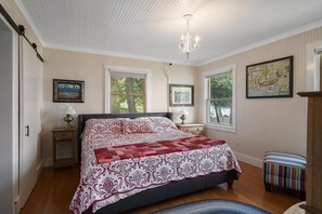 A bedroom with a large bed, two bedside tables, a chandelier, and two windows. - Charming Burt Lake Cottage (Cheboygan)