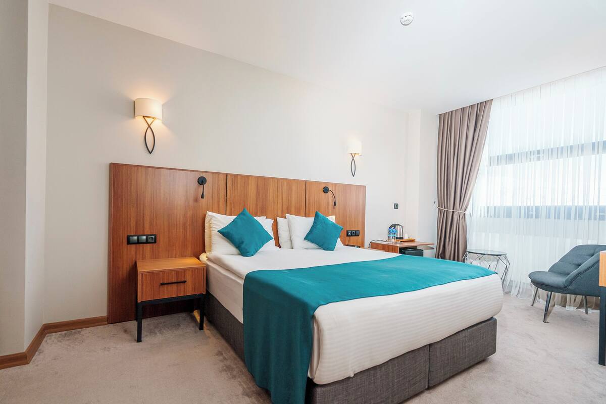 Quarto de hotel com uma cama grande, duas mesas de cabeceira, uma cadeira, uma mesinha e uma janela com cortinas.