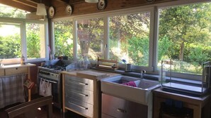 Een keuken met houten balken, een gasfornuis, een spoelbak en grote ramen met uitzicht op een tuin.