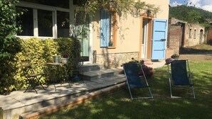 Een gebouw met een beige gevel, blauwe luiken en een stenen trap die naar een deur leidt. Twee blauwe stoelen staan buiten op een gazon.