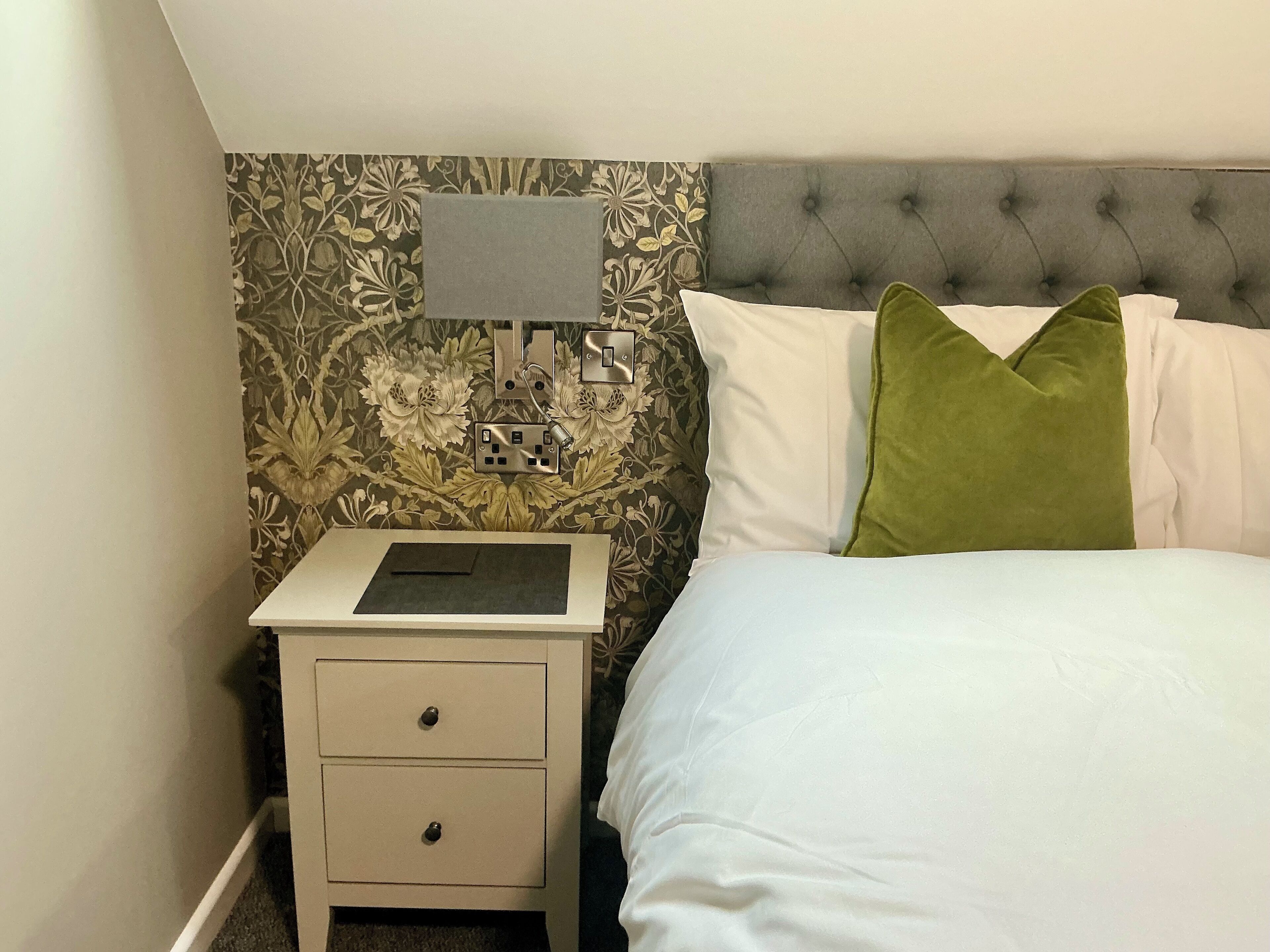 A bedroom with a bed, a nightstand with drawers, a lamp, and a wall with floral wallpaper.