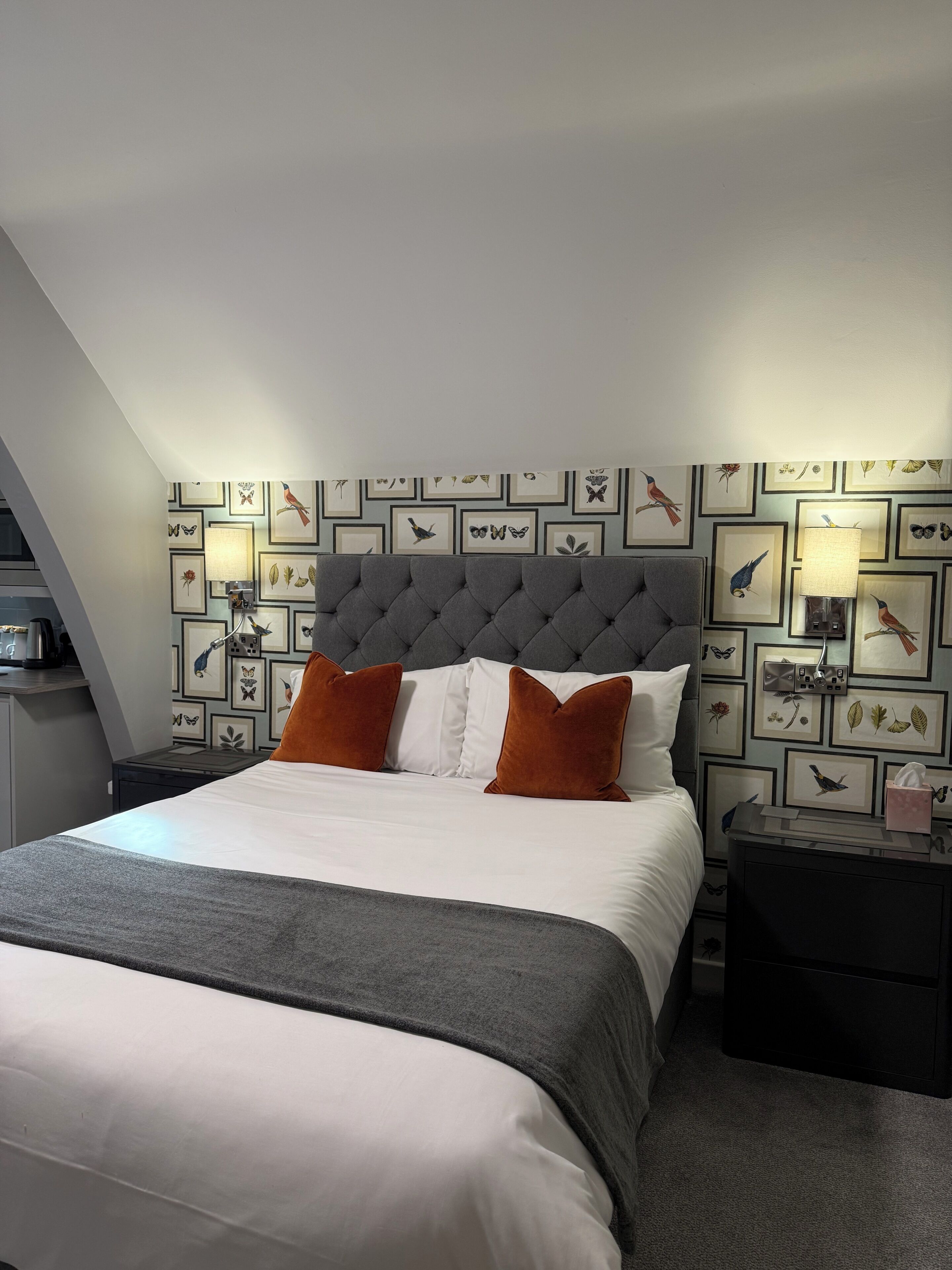 A neatly made bed with a grey headboard, white and orange pillows, and a grey throw blanket. There are framed pictures on the wall, a bedside lamp, and a small table with a tissue box.