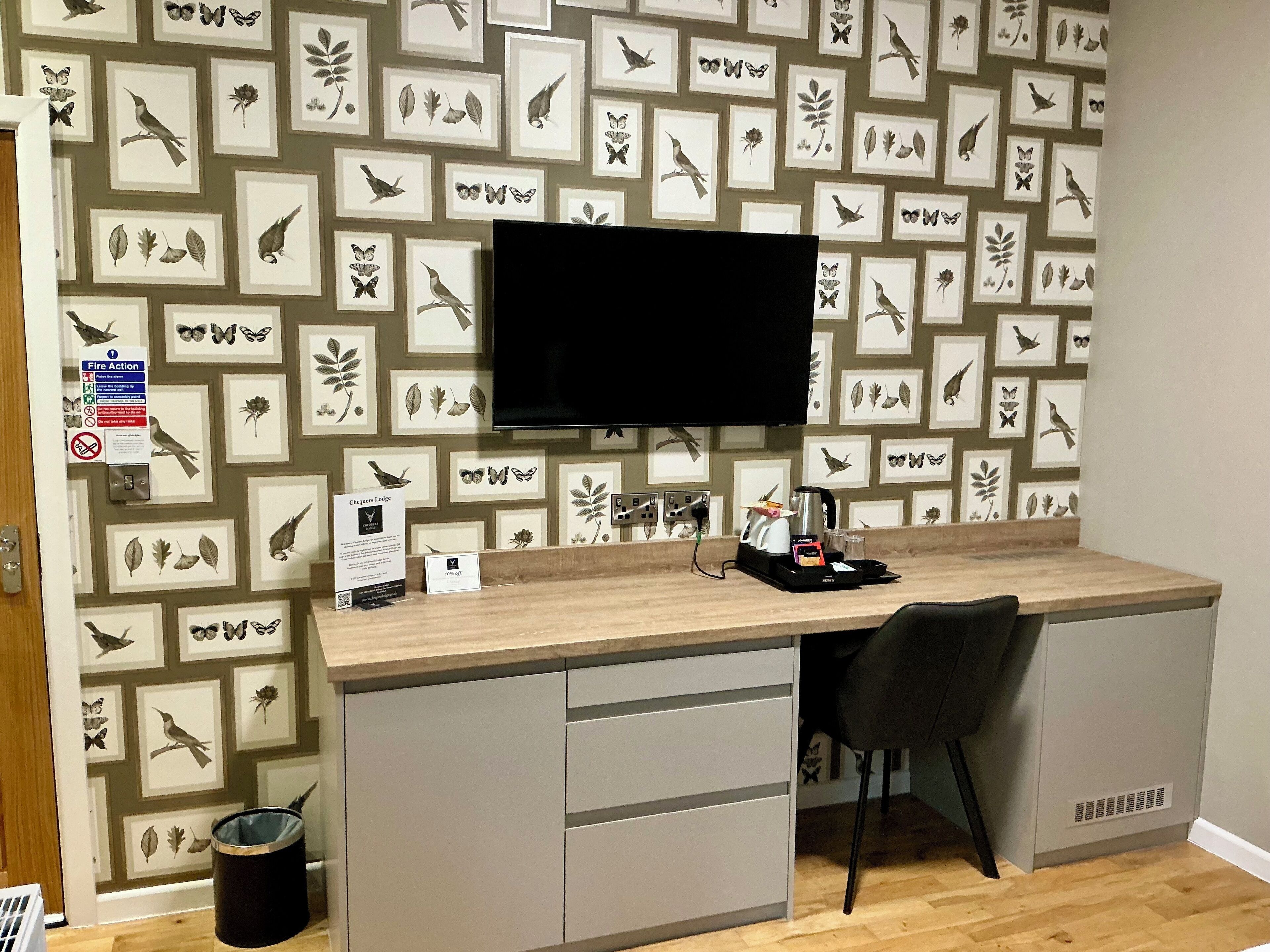 A room with a wooden desk, a black chair, a flat-screen TV mounted on the wall, and a wall decorated with framed nature prints.