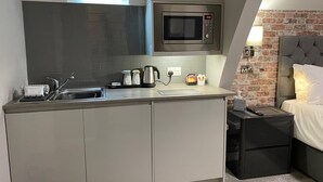 Una cocina compacta con microondas, hervidor de agua y cafetera. Una mesita de noche con control remoto está junto a una cama con cabecera.
