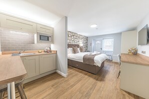 A compact hotel room with a kitchenette, a bed with a headboard, a small desk, and a TV mounted on the wall.