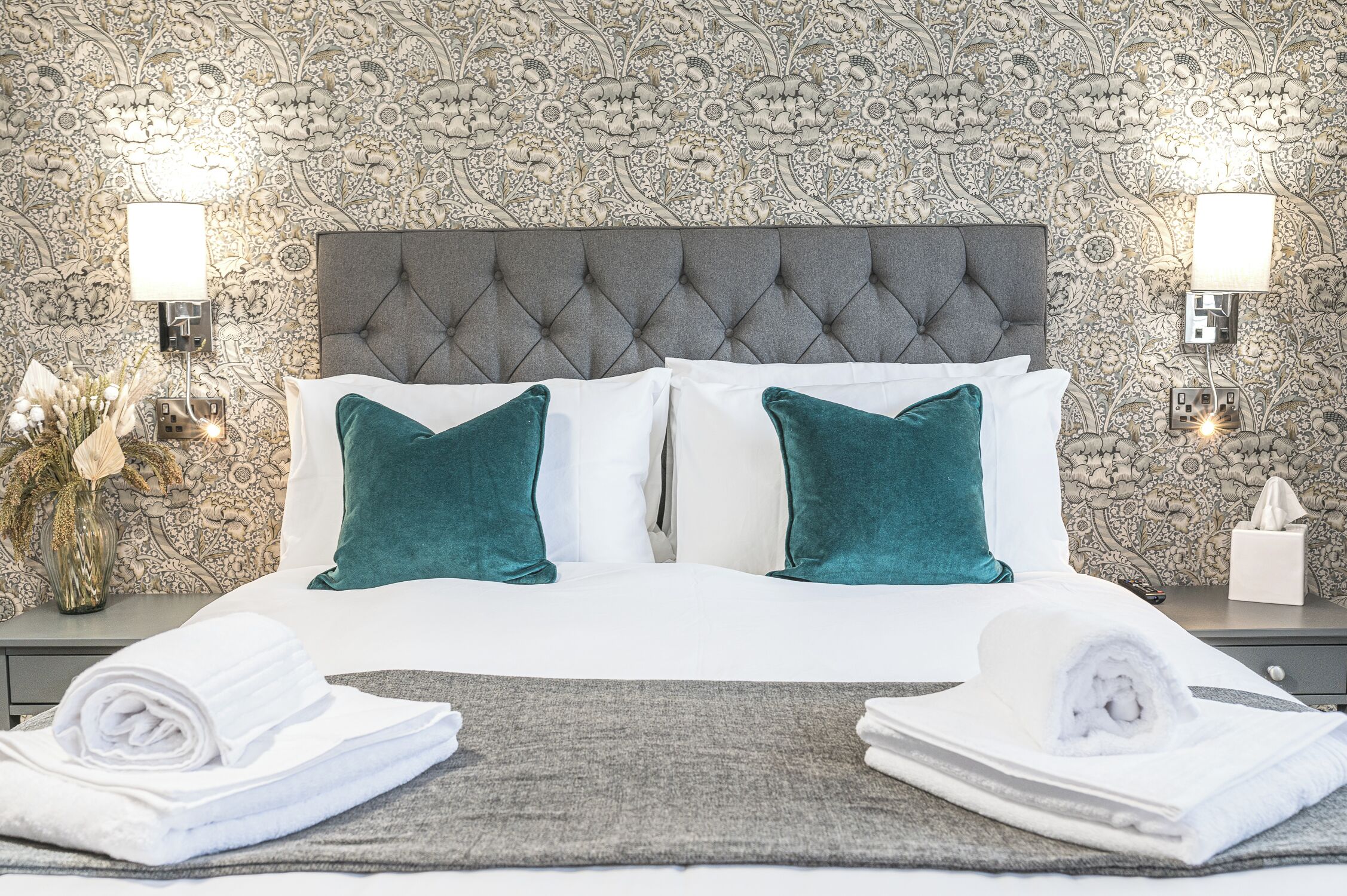 A neatly made bed with a tufted headboard, two teal pillows, and white linens. There are two bedside lamps, a vase with dried flowers, and a tissue box on the nightstand.