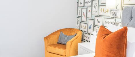 A hotel room with a white bed, a small orange chair, and a wall decorated with framed botanical prints.
