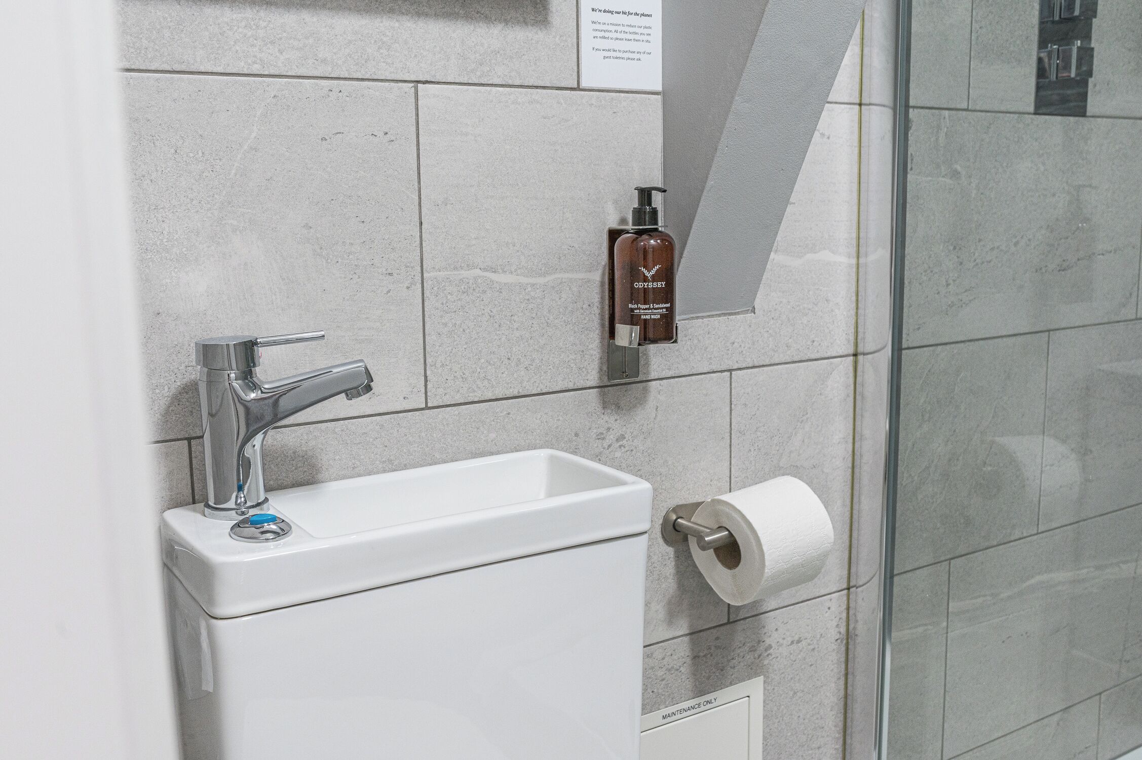 Een badkamer met een witte wastafel, een zilveren kraan, een fles handzeep en een toiletrol.