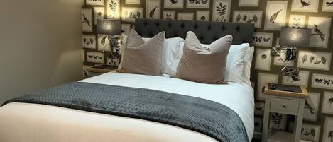 A neatly arranged bedroom with a bed, pillows, a bedside table, and a wall with framed botanical prints.