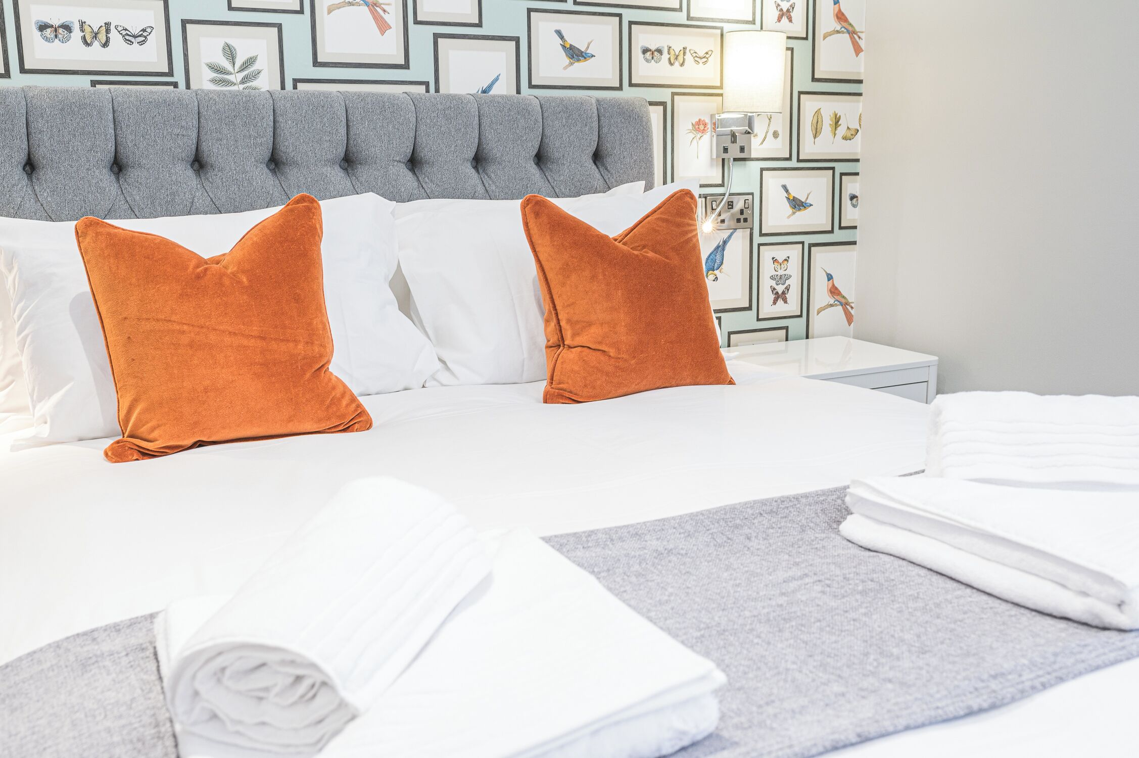 Ein ordentlich bezogenes Bett mit weißen Leinen und zwei orangefarbenen Kissen, ein grauer möbelschoners Kopfseitenverkleidung und eine Wand mit gerahmten botanischen und Vogelzeichnungen.