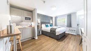 Una habitación de hotel moderna con cocina, una cama con cabecera gris y una ventana con cortinas.