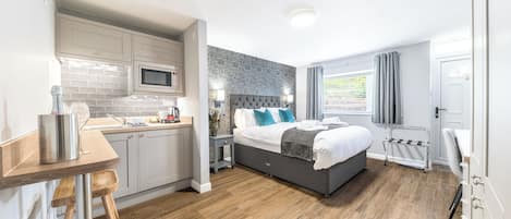 A modern hotel room with a kitchen area, a bed with a grey headboard, and a window with curtains.
