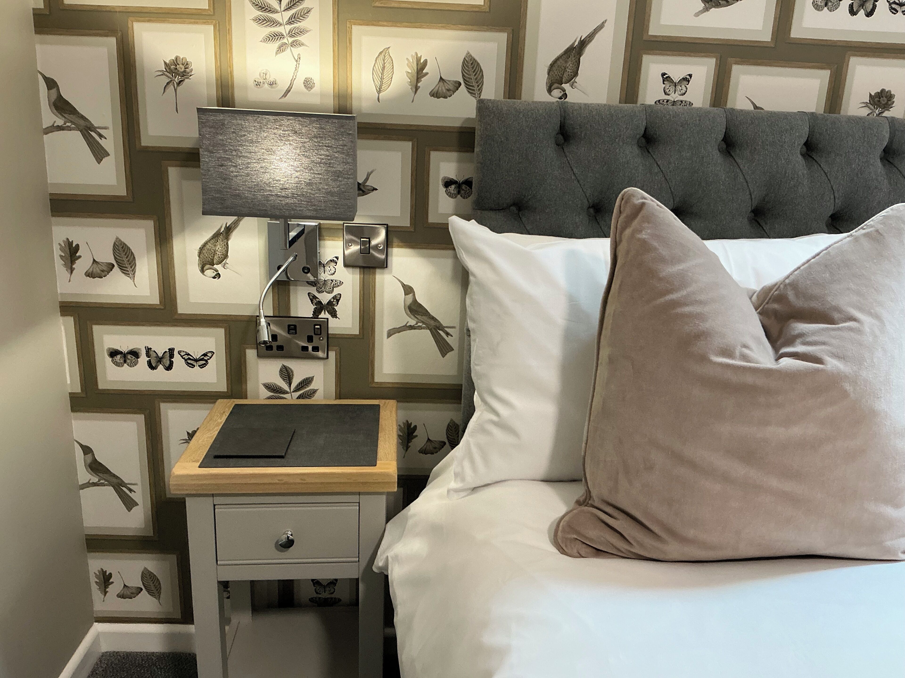 Une chambre à coucher avec une tête de lit capitonnée grise, du linge de lit blanc et une table de chevet avec une lampe.