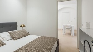 Une chambre moderne avec un lit, une table de chevet, une lampe et une vue sur un coin repas, visible à travers une porte ouverte.