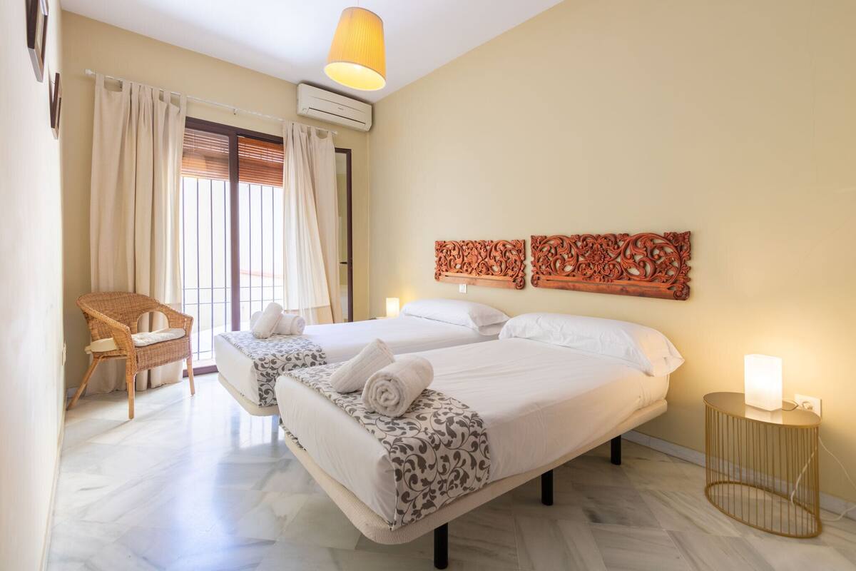 A hotel room with two beds, a wicker chair, a wall-mounted air conditioner, and a bedside table with a lamp.