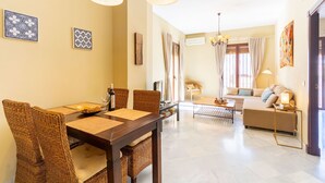 Un comedor con una mesa de madera y sillas de mimbre, un sofá y una lámpara en una sala de estar con piso y paredes de color claro.