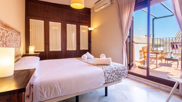 Habitación de hotel con una cama grande, cabecera de madera, balcón con mobiliario exterior y aire acondicionado.