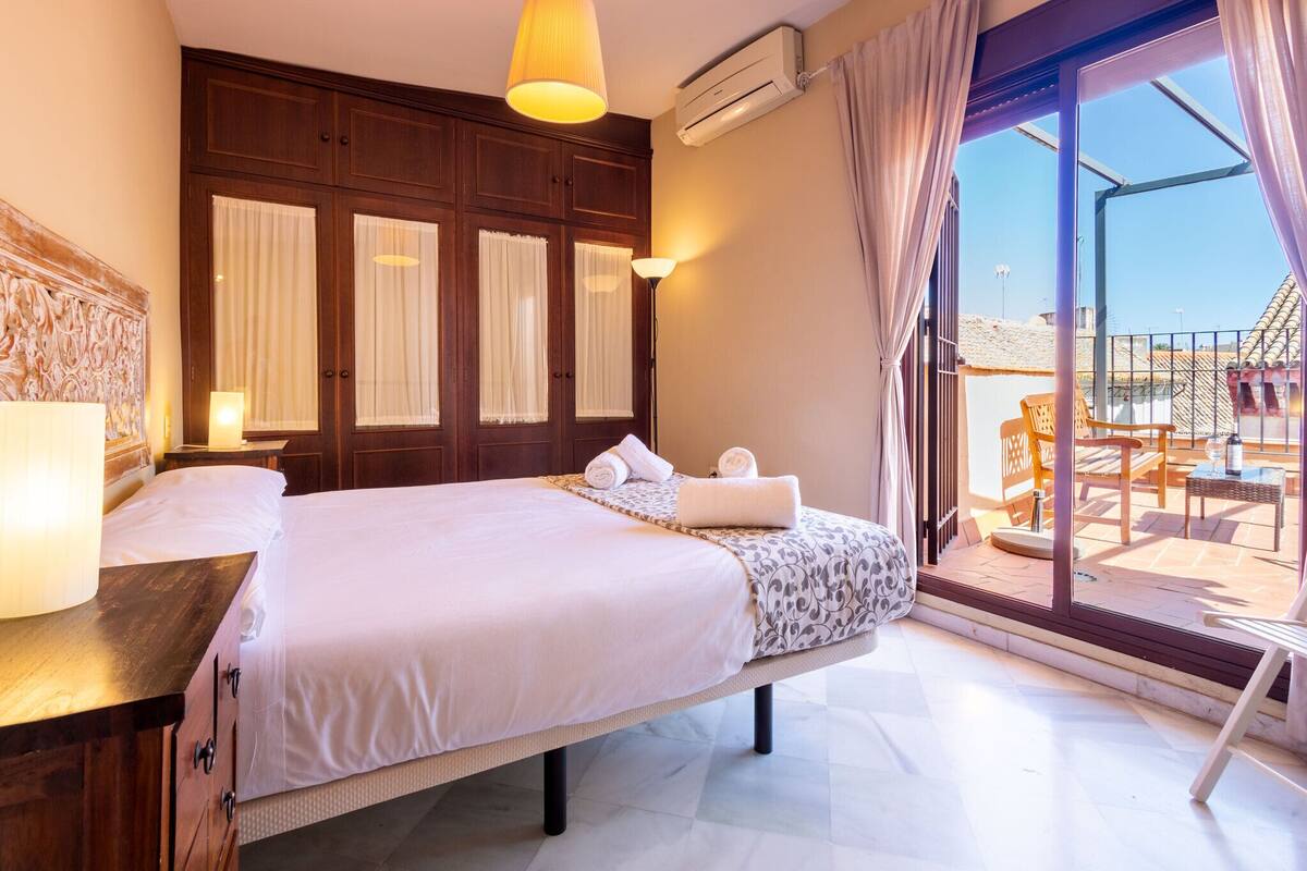 A hotel room with a large bed, a wooden headboard, a balcony with outdoor furniture, and an air conditioning unit.