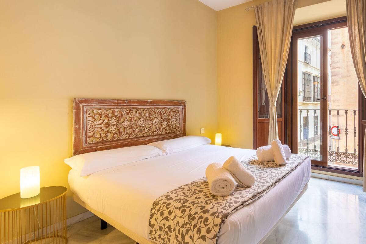 A hotel room with a large bed, a wooden headboard, a balcony with a view, and a round side table with a lamp.