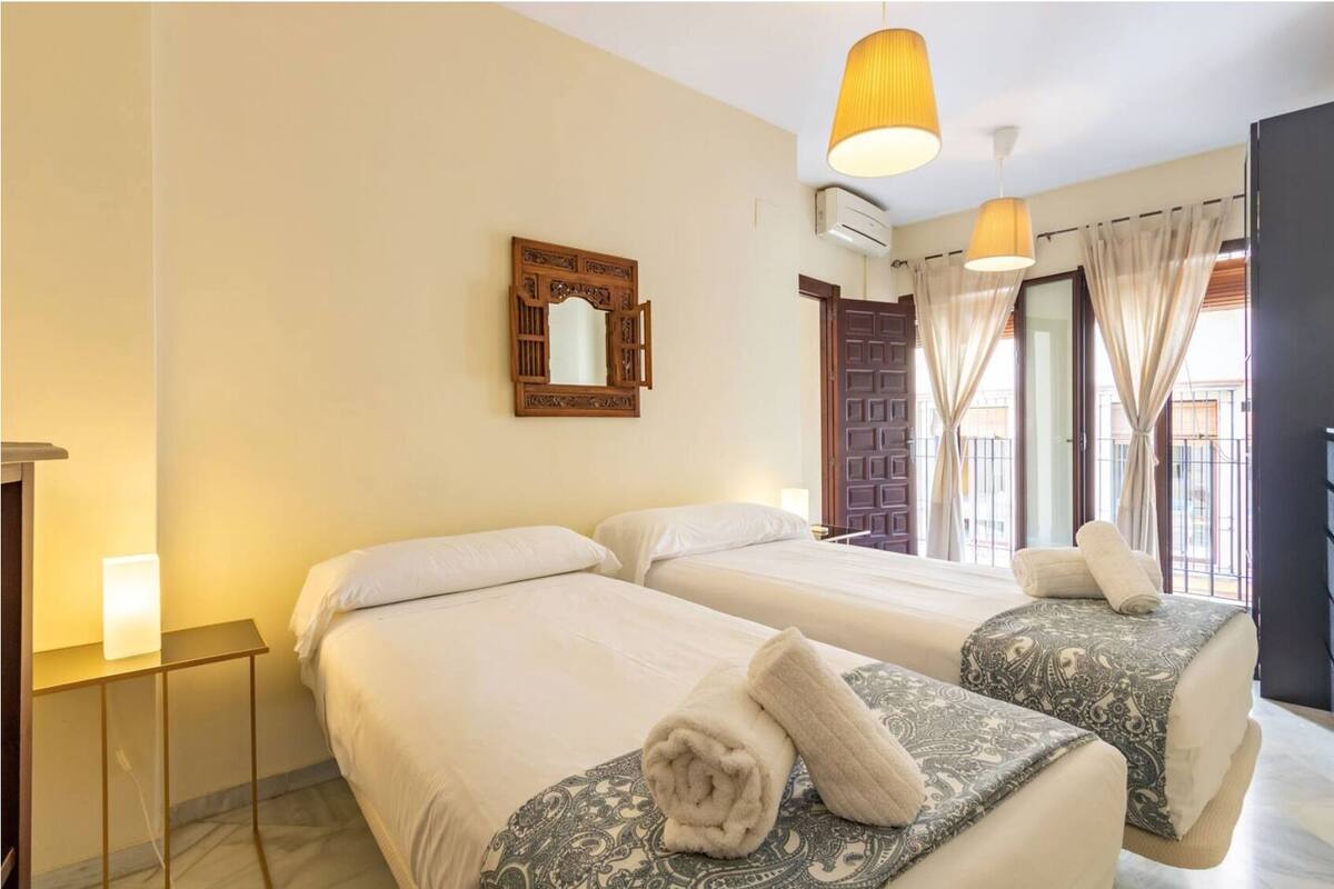 A hotel room with two beds, a wooden-framed mirror, a small bedside table with a lamp, and a large window with curtains.