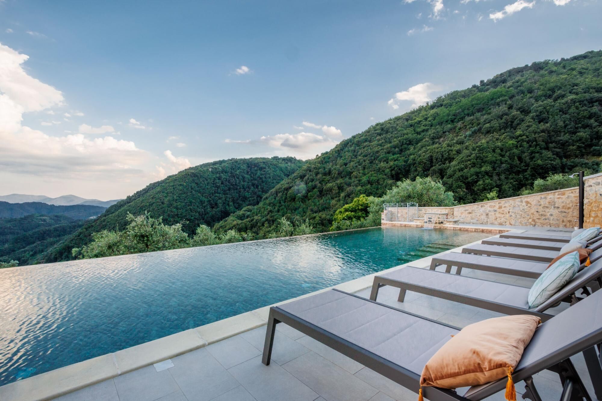 A infinity pool with lounge chairs and a stone wall, overlooking a forested hillside.
