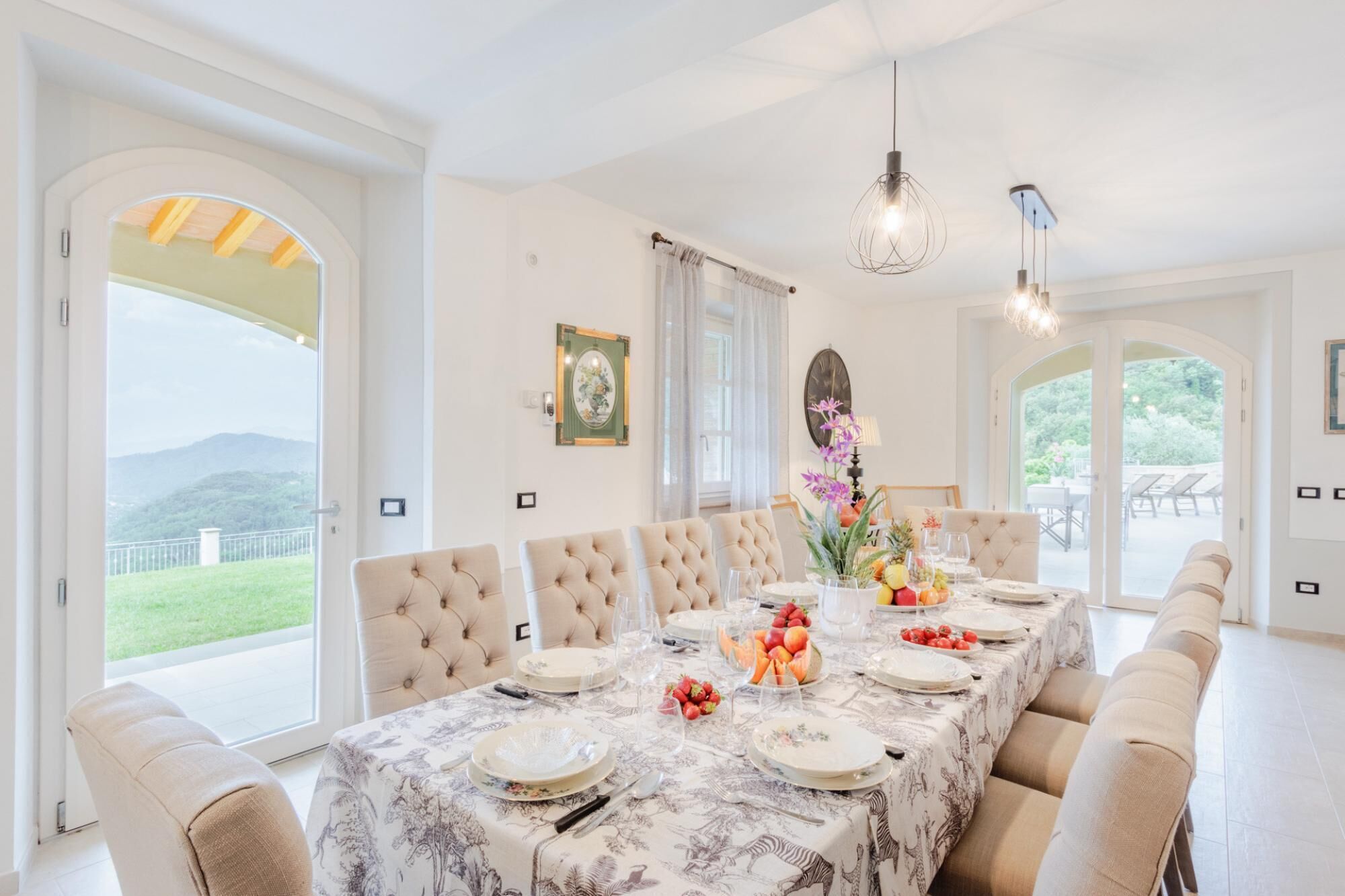 A dining room with a large table set for six, surrounded by six upholstered chairs. The table is covered with a floral tablecloth, and there are plates, glasses, and a fruit centerpiece. The room has large windows with a view of greenery and mountains.