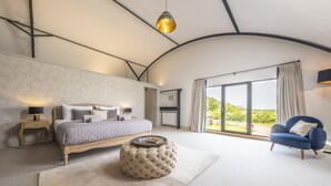 Een slaapkamer met een bed, nachtkastjes, een rond poefje, een blauwe fauteuil en een groot raam met uitzicht.